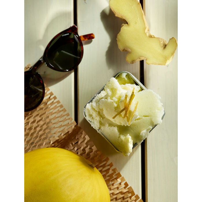 Das Unold Eisbuch โ 101 Eisrezepte zeigt einen Metallbehรคlter mit hellgrรผnem Eis mit Ingwerstreifen, der auf einem weiรen Holztisch neben frischem Ingwer, gelber Melone und einer Sonnenbrille steht.