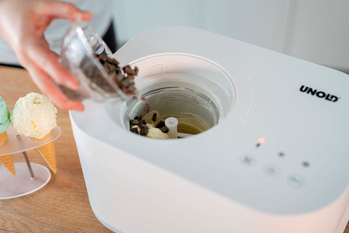 Eine Person füllt Schokoladenstückchen in eine mit Vanilleeis gefüllte Unold Eismaschine Snow 1 L. Daneben stehen drei Waffeln mit grünen und gelben Kugeln auf einem Teller. Die weiße Maschine trägt die Marke Unold.