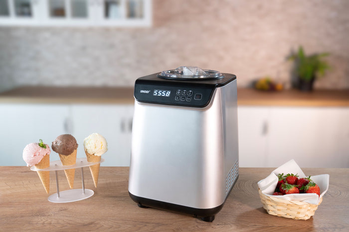 Die Unold Eismaschine Uno 1,2 L steht auf einer hรถlzernen Kรผchentheke neben einem Stรคnder mit drei Eistรผten und einem Korb Erdbeeren. Ihre Digitalanzeige zeigt 55:58.