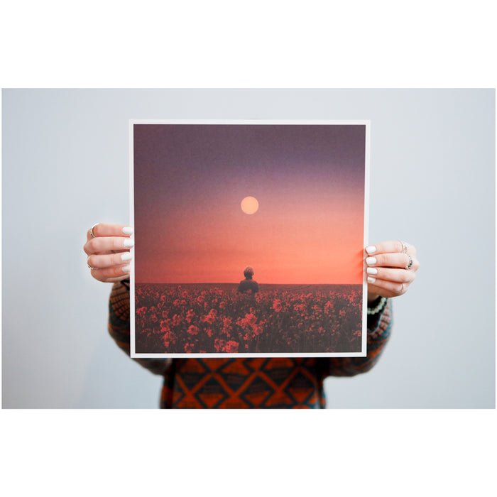 A person holds a Brent Walsh lithograph poster featuring a silhouette standing beneath a vibrant sun in an orange and purple sky over a flower-filled field at sunrise or sunset.