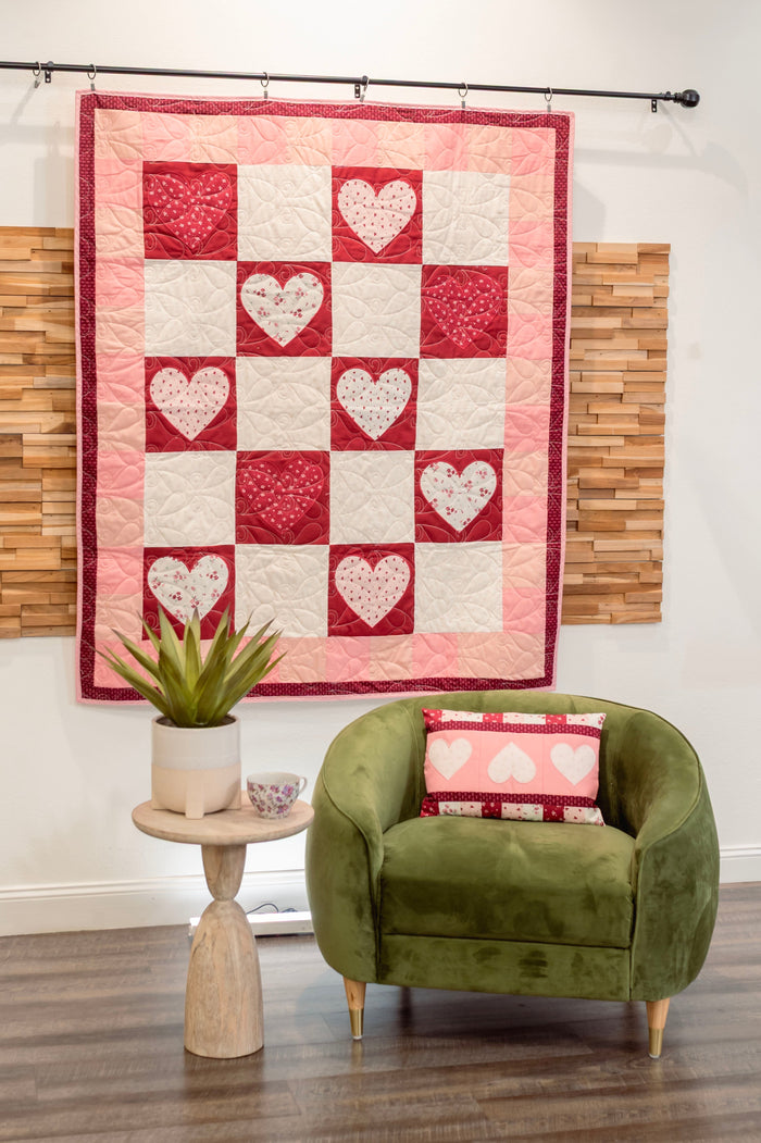 Quilt with heart pattern hanging on a wall above a green armchair with a matching pillow.