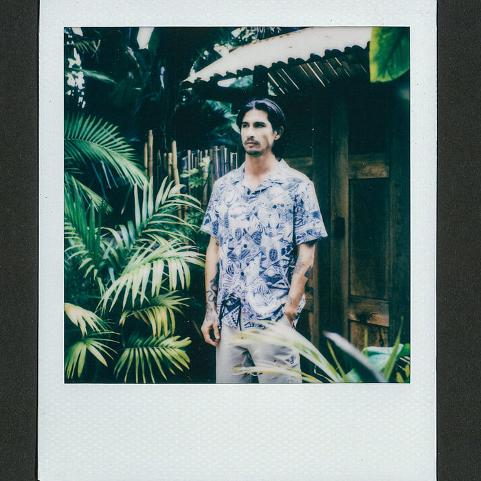 Man wearing a blue patterned Beckham Camp Shirt standing outdoors by tropical plants and wooden structure in relaxed pose