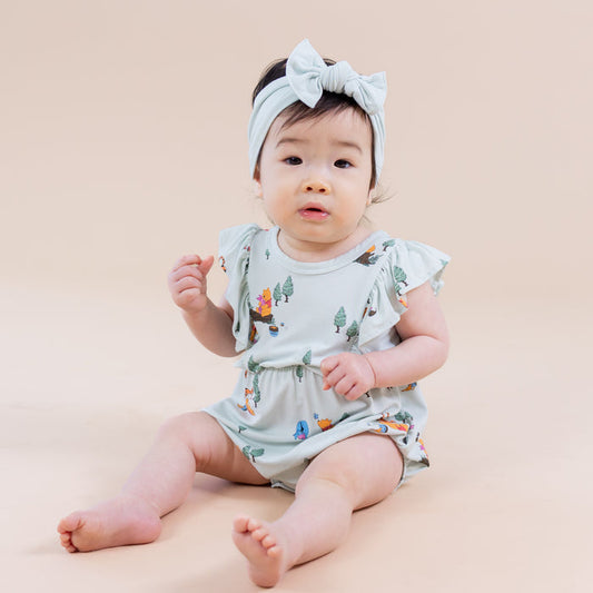 Young toddler sitting on the floor wearing the Bubble Romper in Winnie the Pooh Hundred Acre Wood paired with a light green bow