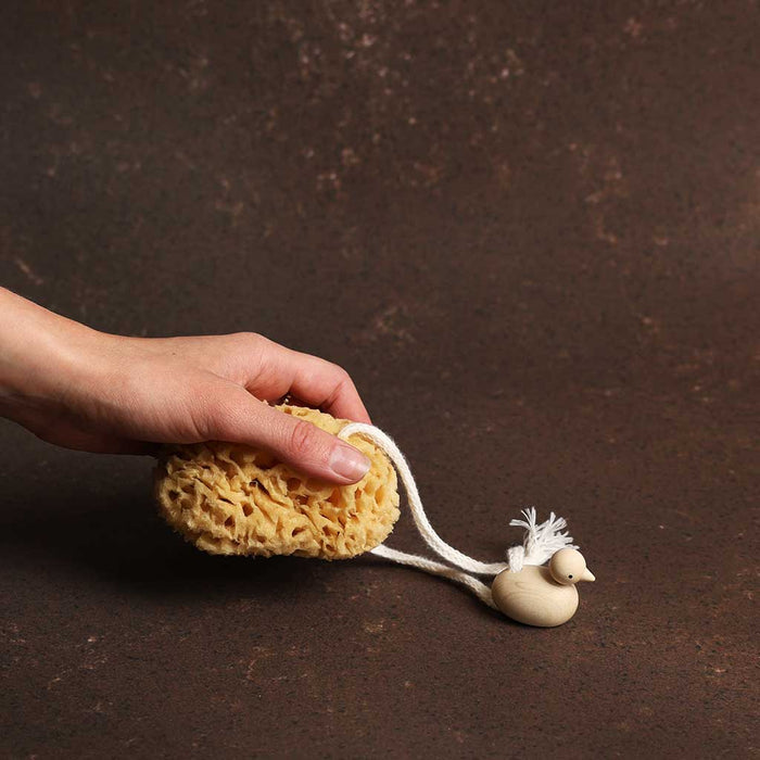 Hand holding a yellow sponge with a white string and a small beige duck toy on a dark brown background