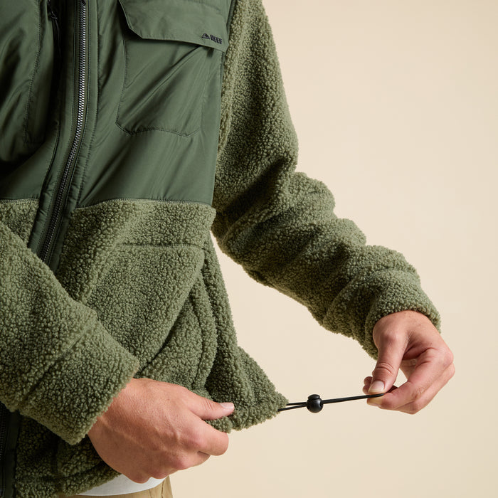 Close-up of hands adjusting the hem drawcord on the olive green Cooke Sherpa Jacket with textured fabric front panel.