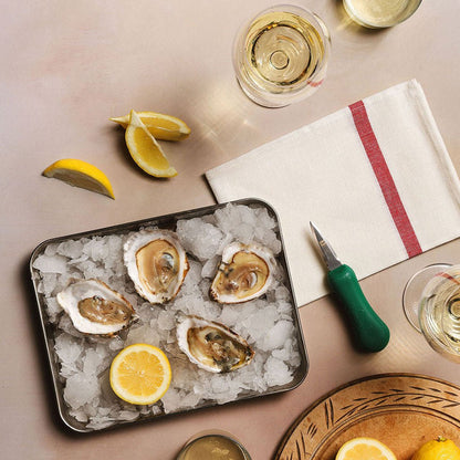Oysters on ice next to shucker and lemon wedges and wine glasses on a table.