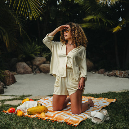 Woman wearing Ellie Short kneeling on a beach towel with side-facing white REEF sandals beside her outdoors