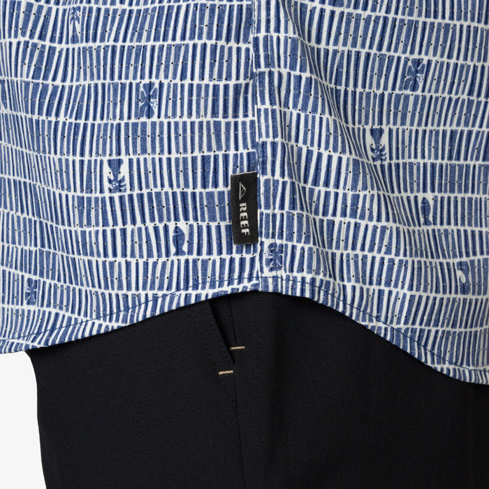 Close-up of blue patterned Ferris Classic Shirt hem with REEF tag over black pants, beach-inspired men's apparel.