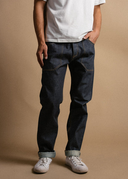 Man in white t-shirt and dark jeans stands casually, hands in pockets, wearing white sneakers against a neutral background.