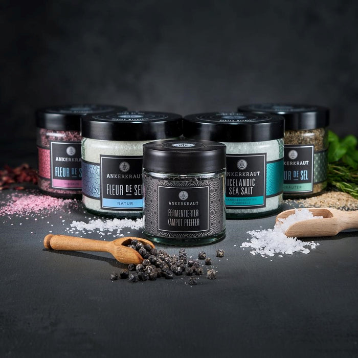 Gourmet Set mit hölzernen Gewürzlöffeln voller Fleur de Sel und Preffer vor schwarzem Hintergrund.