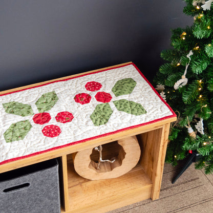 Decorative table runner with holly design on a wooden bench next to a Christmas tree.
