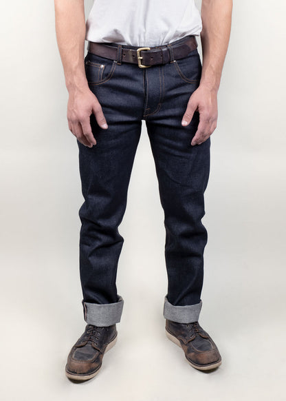 Man in dark jeans and brown leather shoes stands against a plain background, showcasing casual style with a white shirt.