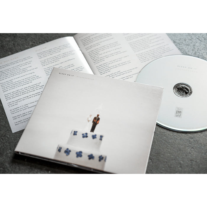 CD with artwork of a weeding cage where the bride and the groom on top of the cake are on fire. The CD itself and the Book with lyrics information are laying beside the case.