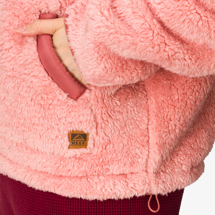 Close-up of a woman’s hand in the front pocket of a coral pink Pilot Sherpa Jacket showing soft, cozy texture and REEF logo.