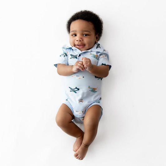 Smiling infant laying on a white surface wearing the Polo Bodysuit in Take Off