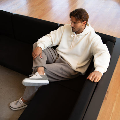 Man sitting on black couch wearing white hoodie and gray pants with Rambler shoes facing left, highlighting comfort and style.