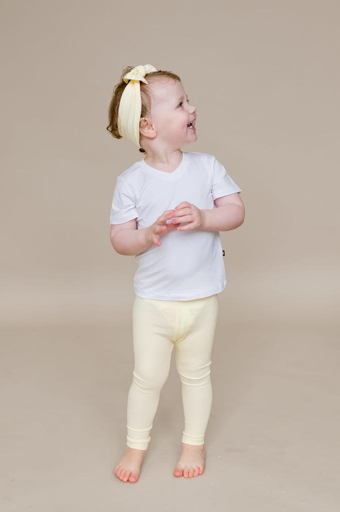 Smiling toddler looking off to the side wearing the Ribbed Leggings in Chamomile paired with a white v-neck shirt