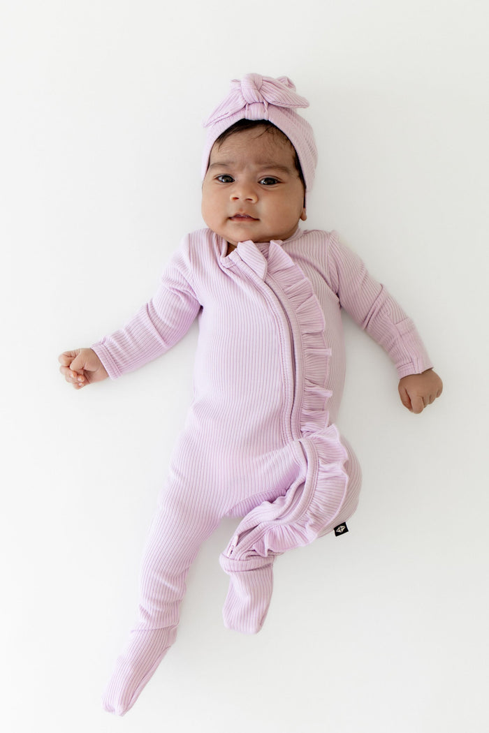 Infant laying on a white surface wearing the Ribbed Ruffle Zipper Footie in Thistle paired with a matching headwrap