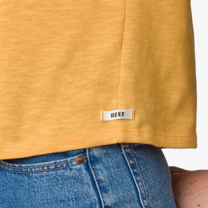 Close-up of the bottom hem of the Ringer Tee in honey gold showing soft fabric and REEF label above blue denim jeans.