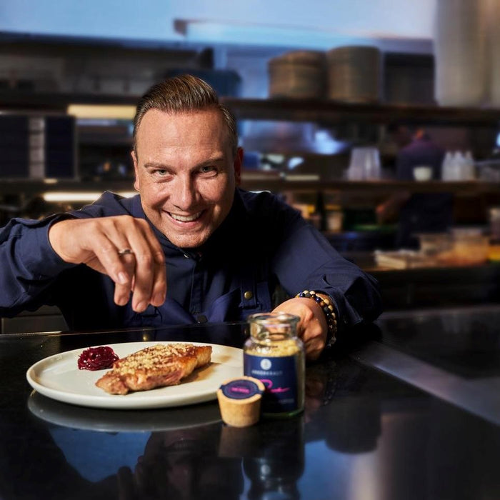 Tim Raue blickt in die Kamera und streut Szechuan Gewürzmischung über ein fertiges Steak vor ihm.