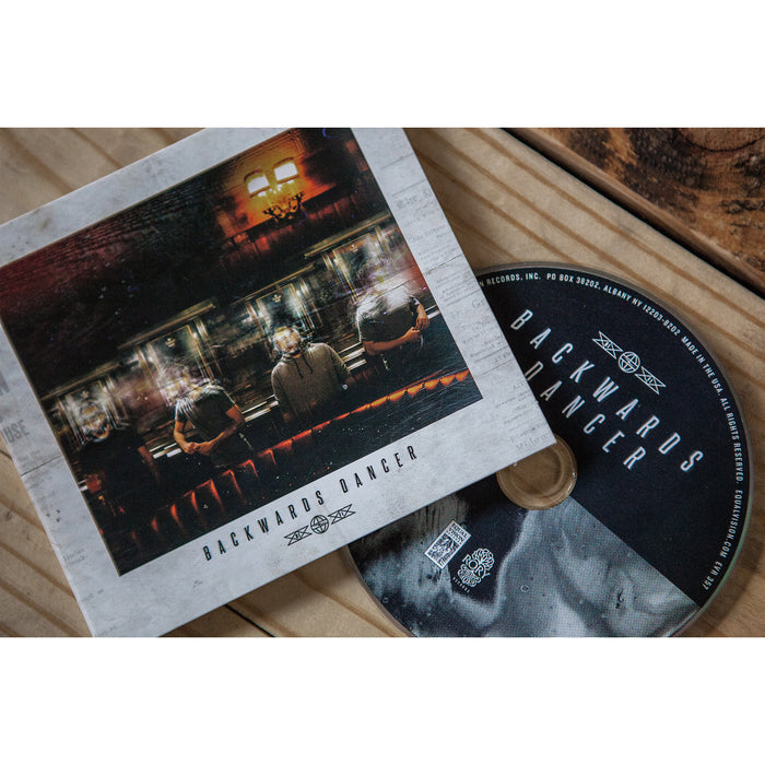 A Backwards Dancer CD and its cover sit on a wooden surface. The cover features three people sitting in a dimly lit room, viewed from behind, evoking an Airy Breathe vibe. The CD is partially pulled from its case.
