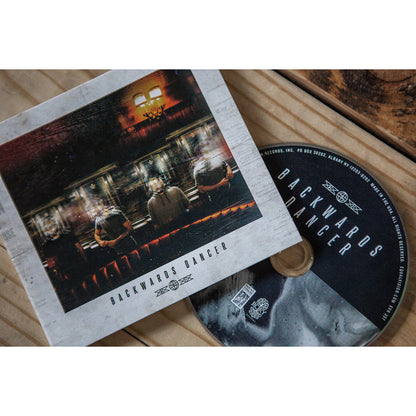 A Backwards Dancer CD and its cover sit on a wooden surface. The cover features three people sitting in a dimly lit room, viewed from behind, evoking an Airy Breathe vibe. The CD is partially pulled from its case.