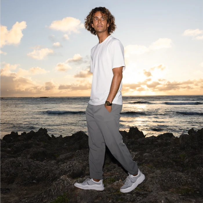 Man wearing Shorebreak TRV shoes facing left in grey walking pants, standing on rocky beach at sunset with ocean background.