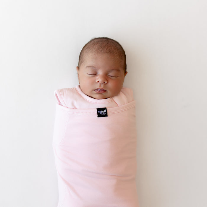 Baby sleeping on a white surface wearing a sleep bag swaddler in a light pink with a kyte baby logo in center