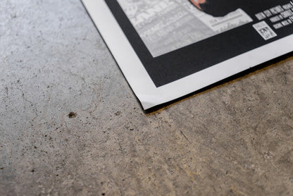 A close-up of a slightly damaged Bane 18x24 poster with a black border on gray concrete. The bottom corner is lifted, revealing text and a QR code, with no refunds faintly visible.