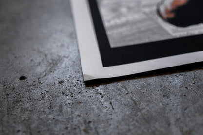 A close-up of a slightly damaged Bane 18x24 poster on textured gray concrete. The edge is slightly bent, and the image features a white and black border.