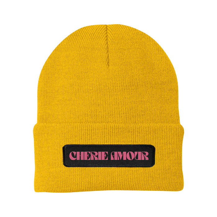 This gold knit beanie by Cherie Amour features a folded brim and a black front patch with “CHERIE AMOUR” in bold, pink, stylized letters.