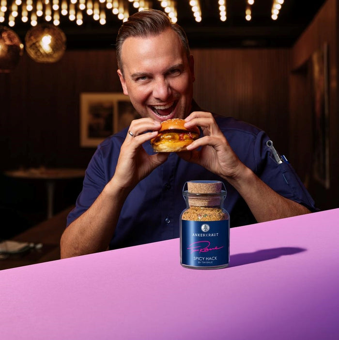 Tim Raue beißt in einen Burger, während vor ihm das Ankerkraut Spicy Hack Gewürz im Korkenglas steht.