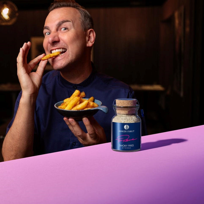 Tim Raue beißt in eine Pommes, während vor ihm das Ankerkraut Smoky Fries Gewürz im Korkenglas steht.