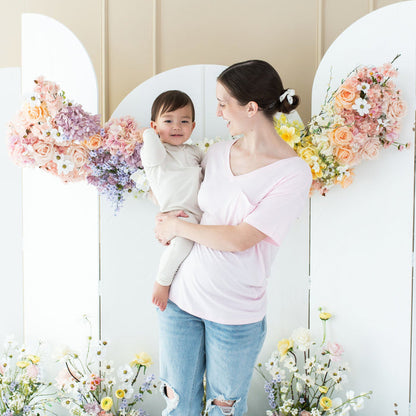 Woman wearing Kyte Baby Women's Relaxed Fit V-Neck tee in Sakura pink holding toddler