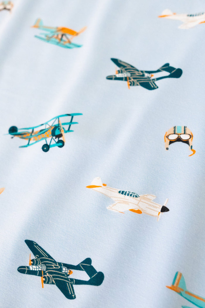A repeating, all-over print featuring: a teal triplane with a light orange checker design; a racing seaplane colored in turquoise and light orange; a night fighter aircraft in teal with light orange accents; a white fighter bomber aircraft with a black nose, teal windows, and light orange accents; and a light grey aviator hat with goggles and blue stripes, over a sky blue background color called Breeze.