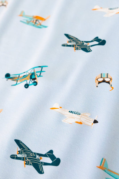 A repeating, all-over print featuring: a teal triplane with a light orange checker design; a racing seaplane colored in turquoise and light orange; a night fighter aircraft in teal with light orange accents; a white fighter bomber aircraft with a black nose, teal windows, and light orange accents; and a light grey aviator hat with goggles and blue stripes, over a sky blue background color called Breeze.