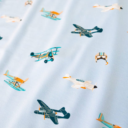 A repeating, all-over print featuring: a teal triplane with a light orange checker design; a racing seaplane colored in turquoise and light orange; a night fighter aircraft in teal with light orange accents; a white fighter bomber aircraft with a black nose, teal windows, and light orange accents; and a light grey aviator hat with goggles and blue stripes, over a sky blue background color called Breeze.